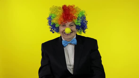 Senior Old Woman Clown in Wig Waves Her Hands, Smiling. Yellow Background