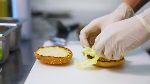 Hands in latex gloves put lettuce on top of fried bun. Making burger on white table.