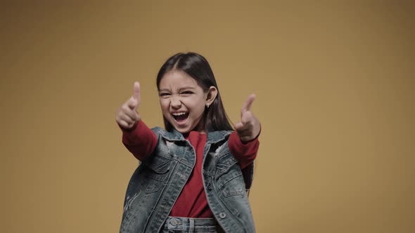 Little Girl Points Her Fingers at the Camera and Smiles on a Yellow Background