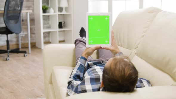 Young Man in Well Lit Living Room Holding a Digital Tablet PC with Green Screen on It