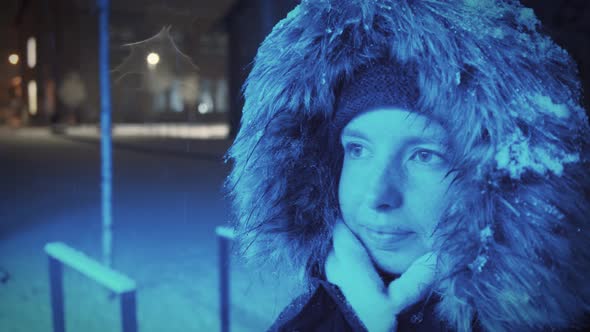 Young woman with a snow-covered fur hood is standing in a blue light in a wintry landscape at night