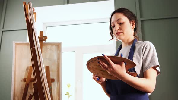 Art Studio  Young Woman Choosing a Brush and Painting on Canvas