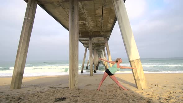 A young attractive woman doing yoga on the beach under a pier.