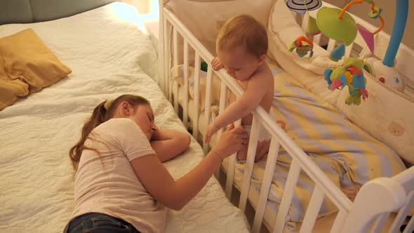 Little Sleepless Baby Boy Rocking in Crib Next to Tired Mother Sleeping in Bed