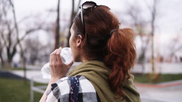 A Girl with a Tail Walks on an Overcast Day in the Park Drinks Water From a Plastic Bottle