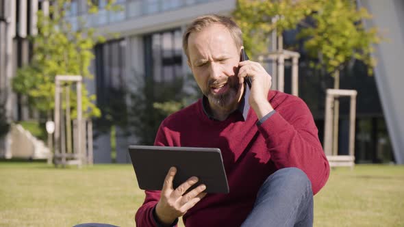 A Middleaged Handsome Caucasian Man Holds a Tablet and Talks on a Smartphone As He Sits in a Street