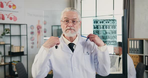 Doctor Putting on His Stethoscope Around His Neck Looking Into Camera in Modern Hospital Office