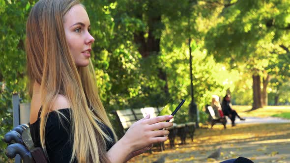 Young Beautiful Woman Sits on the Bench in Park and Works on the Tablet