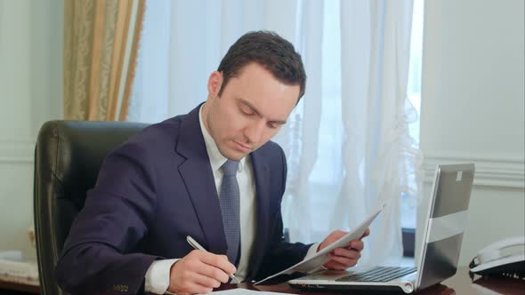 A Young Attractive Businessman Working at His Desk, Takes a Phone Call, Makes Notations, Drinking