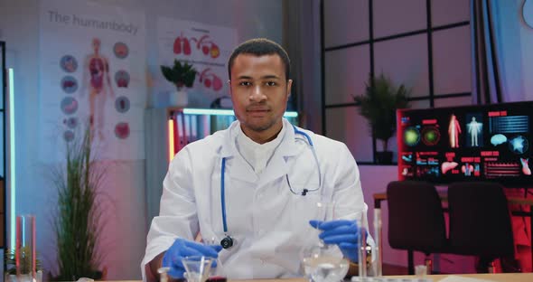 African American Chemist in white Coat posing on camera with glass flask and test tubes filling