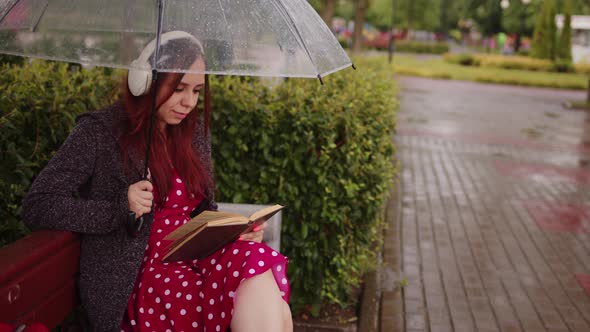 Young Beautiful Woman in Headphones with Transparent Umbrella Reading Book Sitting on Bench in City