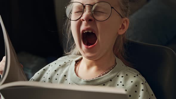 Little child tired girl reading a book in glasses after school and yawns