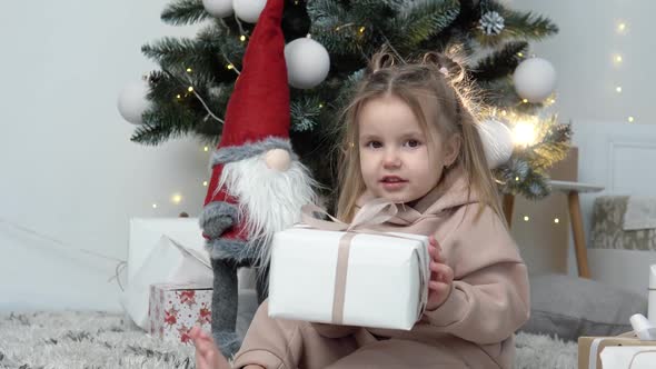 Sweet Little Girl Sitting Near Christmas Tree with a Present Box in Her Hands