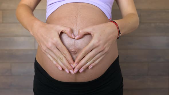 Pregnant woman forms a heart with hands on big belly button, close up