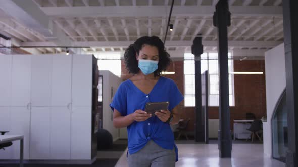 Mixed race businesswoman walking through corridor wearing facemask holding tablet