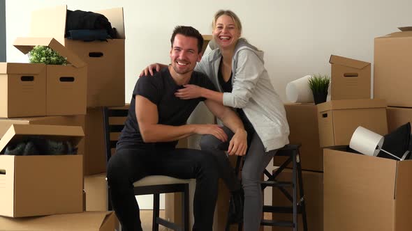 A Happy Moving Couple Sits on Chairs, Looks at the Camera and Celebrates in an Empty Apartment