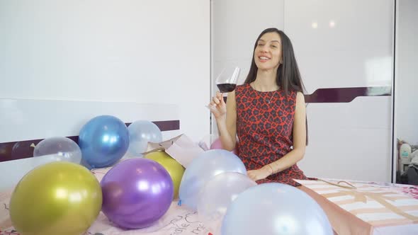 Beautiful Birthday Woman with Red Dress Sitting on Bed with Balloons and Holding a Glass of Red Wine