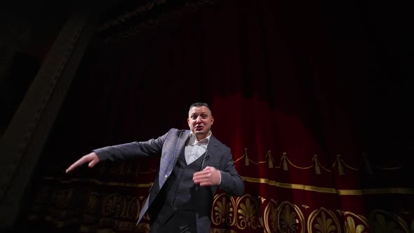 Senior male entertainer dances on stage in front of theater hall. Speaker akes audience laugh.