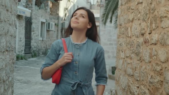 Pretty young woman with brunette hair in denim overalls walks and smiles in old town in Montenegro
