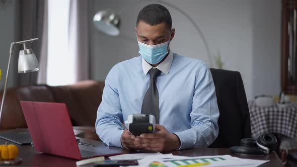 Focused African American Man in Covid19 Face Mask Using Label Maker and Typing on Laptop Keyboard