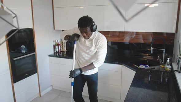 Happy Black Man Listening Music on Headphones Dancing and Singing While Cleaning Floor in the