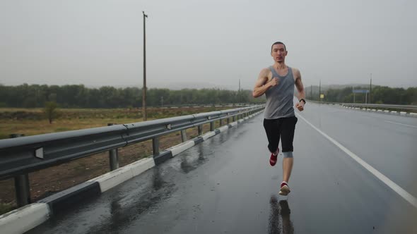 Athletic man runs outdoor on road in steppe doing workout during rain, front view. Man runs along