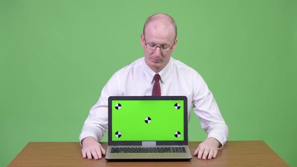 Happy Mature Bald Businessman Showing Laptop Against Wooden Table