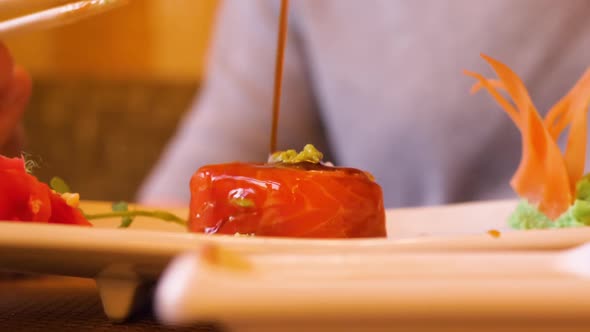 Male Hand Holding Sushi Roll with Chopsticks and Pouring Soy Sauce in Restaurant