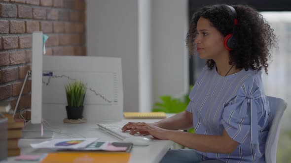 Cheerful Young Woman Listening to Music in Headphones Smiling and Typing on Computer Keyboard