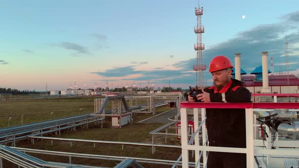 An Employee in a Helmet and Overalls Stands on a Metal Tower of a Gas Tank Makes an Inspection at an