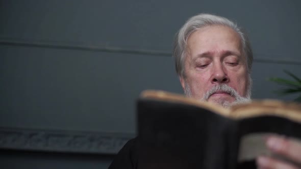 Closeup Lowangle View of Focused Bearded Senior Aged Man Reading Older Paper Book Holding in Hand