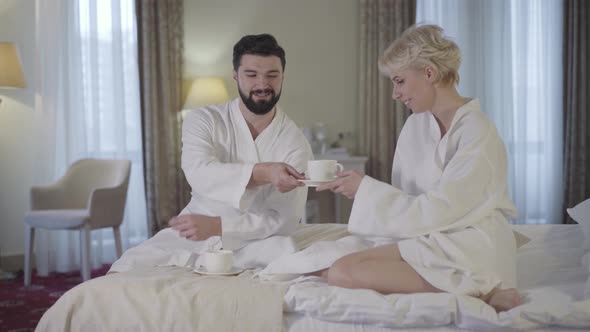 Happy Adult Man and Woman in Bathrobes Clinking Tea Cups in Hotel Room