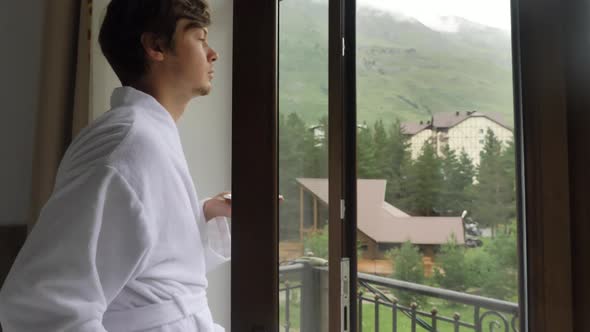 Young Guy Drinks Coffee and Looks at Mountainous Village