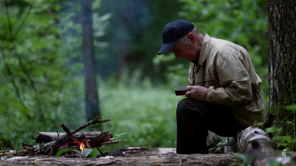 Old Man is Trekking Alone in Forest Resting Near Campfire and Drinking Hot Tea