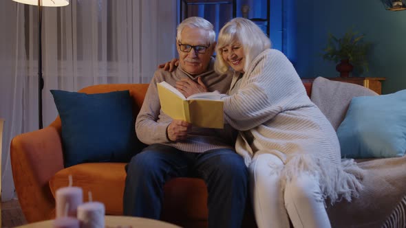 Senior Grandparents Couple Relaxing Reading Book Talking Enjoying Leisure Hobbies at Night Home