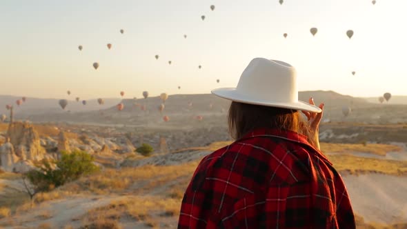 Woman In Hat Walking In Cappadocia. Colorful Hot Air Balloons Fly On Background