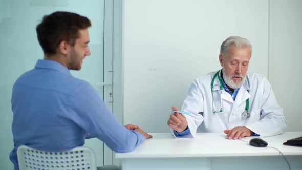 Mature Male Adult Doctor Interviewing Sick Male Patient and Writing Prescription Sitting at Table in