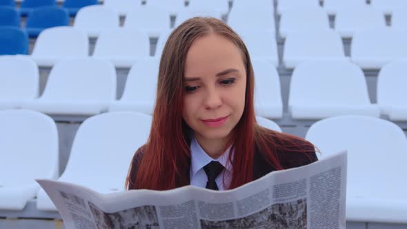 A Woman in a Business Suit Reads a Newspaper While Sitting at the Stadium