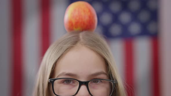 Closeup Eyes of Teen Schoolgirl in Eyeglasses with Apple on Head and American Flag at Background alt