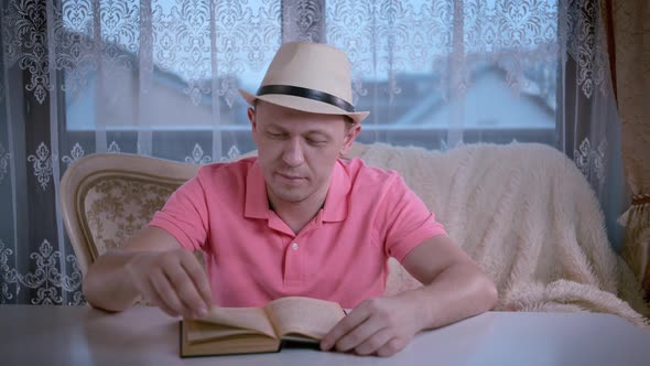 A Man in a Hat in the Evening Sitting at a Table on a Sofa Covered with a Blanket and Reading