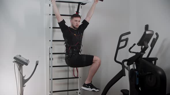 Concentrated Man Wearing Electrostimulation Equipment Hanging By the Stand and Pumping His Lower