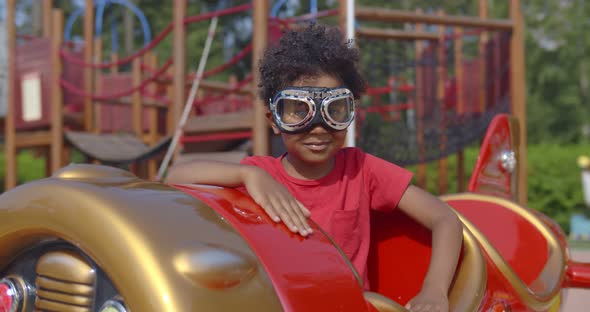 Portrait of Adorable African Toddler Boy Wearing Pilot Goggles Sitting in Toy Plane on Playground