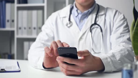 Male Doctor Uses Smartphone While Sitting At His Desk In Hospital. Symptoms And Treatment Of Disease