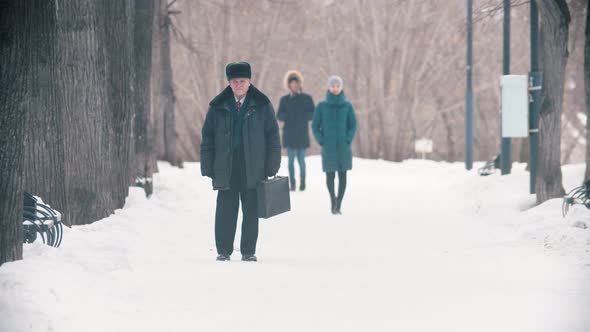 An Elderly Man with Bag Walking in the Snowy Park