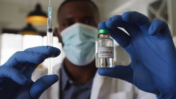 African american doctor wearing face mask holding covid-19 vaccine and syringe at home