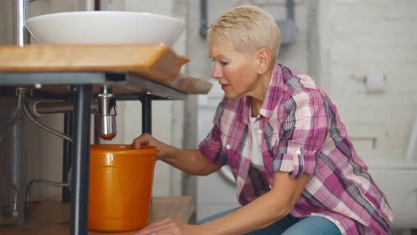 Worried Senior Woman Collecting Water Leaking From Sink in Bucket