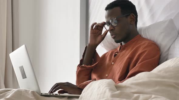 Concentrated AfricanAmerican Man Works on Laptop in Bed