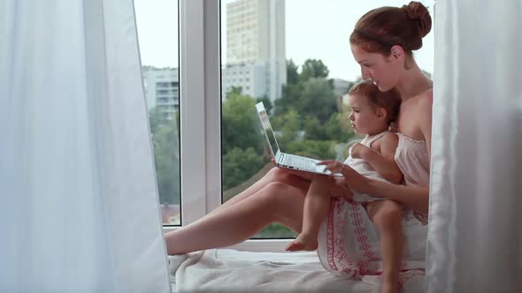 Young Mother and Her Little Daughter with Laptop on the Windowsill