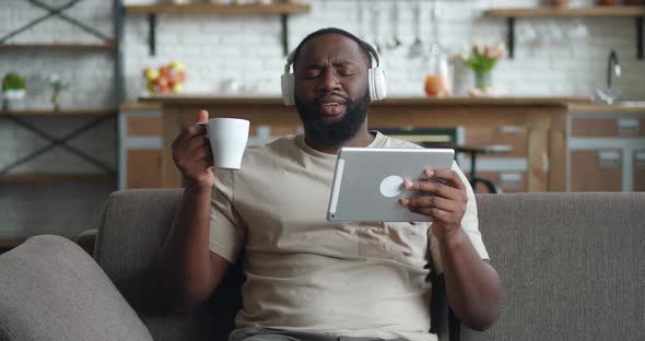 Smiling Black Man in Headphones Using Digital Tablet Sit on Couch at Home Young African Guy Drinking