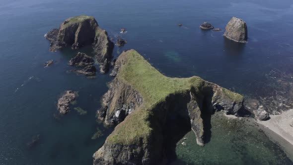 Aerial descends to reveal eroded sea caves in jagged islet in blue sea